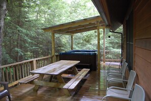 Una terraza de madera con mesas y sillas de picnic, un jacuzzi y una zona cubierta con vistas a un bosque.