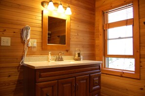Un baño tipo cabaña de madera con tocador de madera, un espejo, un secador de pelo y una ventana con persianas.