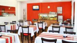 A restaurant with multiple tables and chairs, a buffet counter, and a television screen on the wall. - Arpini Hotel (Rio Grande)