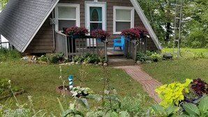 A house with a sloped roof, a porch with a blue chair, and a garden with pink flowers and green foliage.
