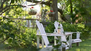 A white outdoor seating area with a table and chairs, surrounded by lush greenery and hanging flower baskets.