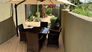 An outdoor patio with a table and chairs under a large umbrella, surrounded by a garden with various plants and a palm tree. - Relaxing Desert Oasis-Salt Water Pool & Jacuzzi/Mountain Views/#067735 3bd (La Quinta)