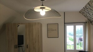 Ein Schlafzimmer mit Bett, einer Pendelleuchte, einem Fenster mit Ausblick, einem gerahmten Bild an der Wand und einer Tür zu einem anderen Raum.