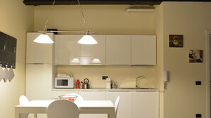 Cozinha moderna com armários brancos, micro-ondas, chaleira e pia. Há uma mesa de jantar com cadeiras e iluminação pendente acima.