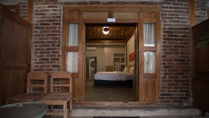 Una habitación rústica con una puerta de madera que da a un dormitorio con cama y ventana. En primer plano, se observa una mesa y una silla de madera.