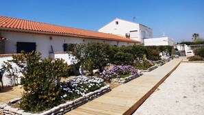 Un edificio blanco con tejado de tejas rojas, rodeado de un jardín con diversas plantas y flores, y un sendero de madera que conduce a la entrada.
