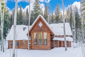 Uma cabana de tronco com telhado triangular, rodeada por árvores cobertas de neve.