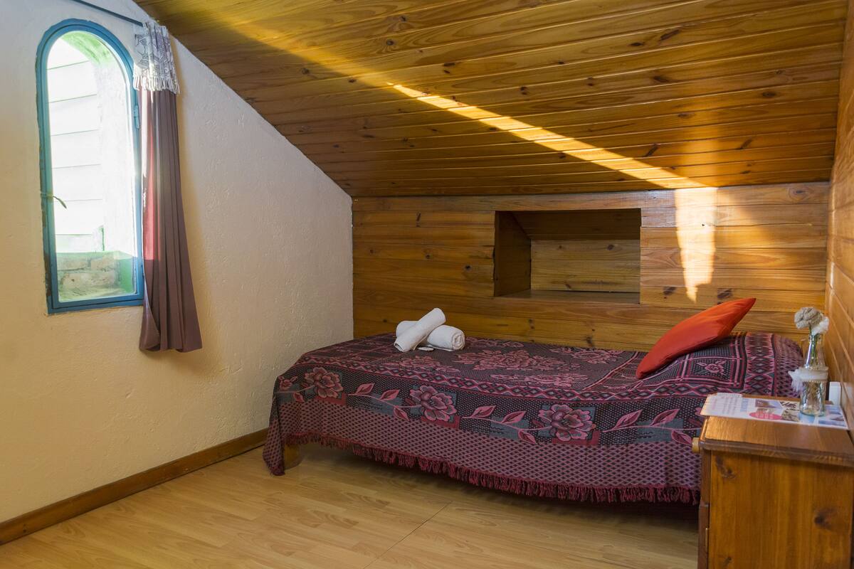 A cozy room with a wooden ceiling, a bed with a patterned blanket, a small window with curtains, and a wooden nightstand with a vase.