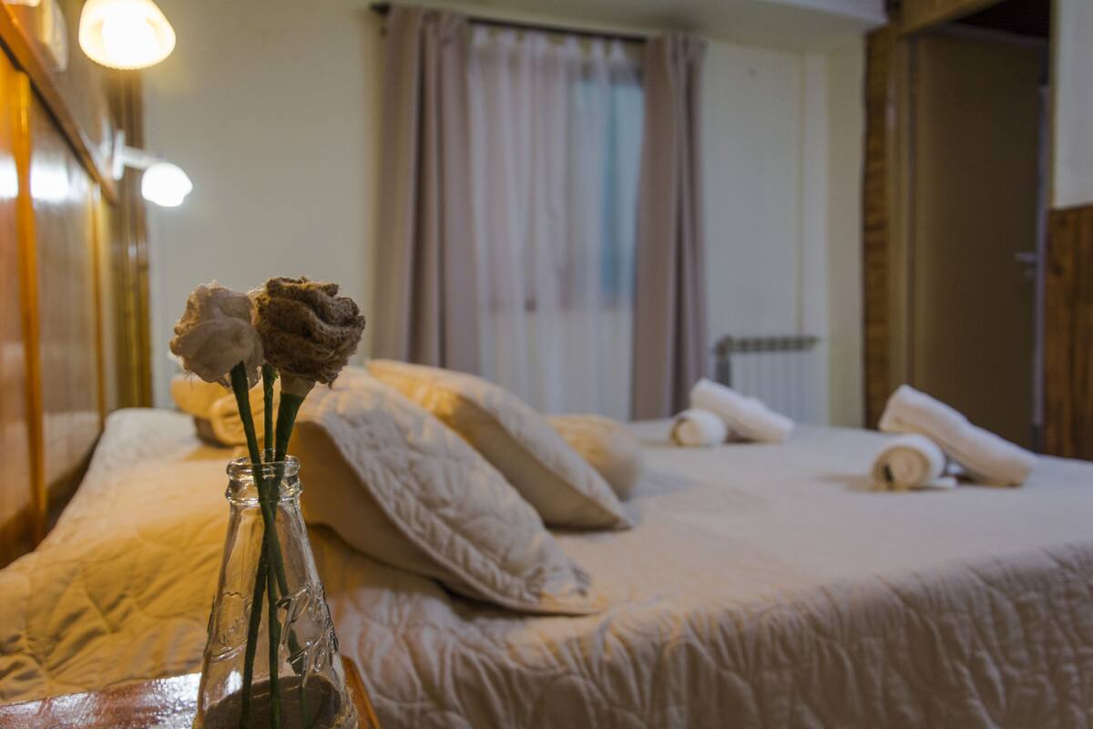 A neatly made bed with pillows and a rolled towel, a vase with flowers on a bedside table, and a window with curtains.