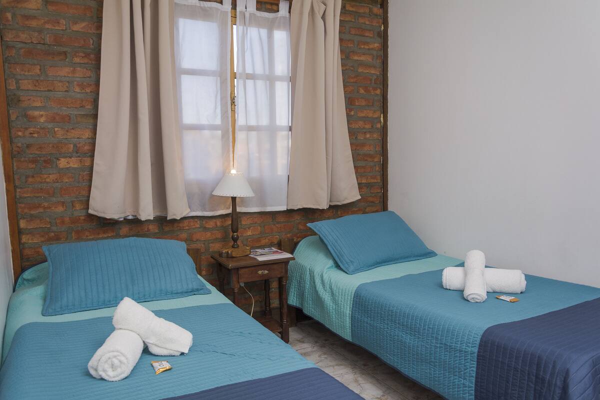 A room with two beds, each with a blue bedspread and pillows, a nightstand with a lamp, and a brick wall.