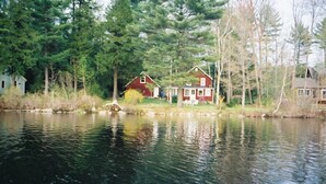 Una cabaña junto a un lago, con fachada roja, rodeada de árboles y una vista despejada del agua.