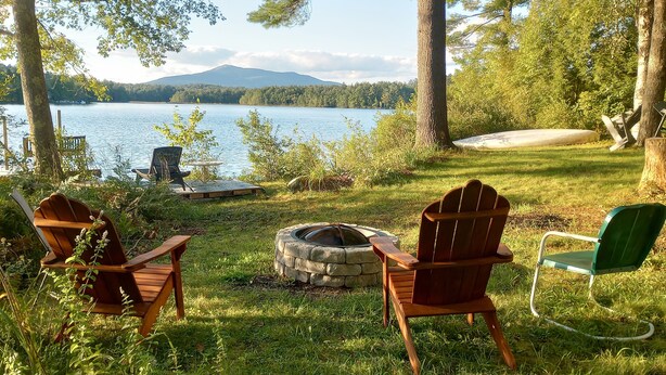 Un espacio al aire libre junto al lago con sillas Adirondack, un hogar de piedra y vistas al lago y las montañas.