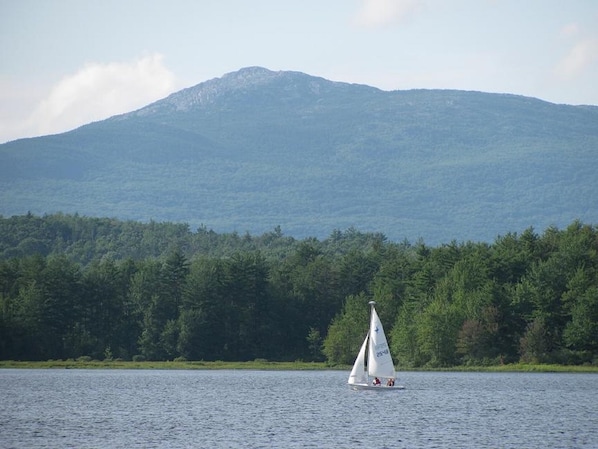 Un velero en un lago tranquilo con una orilla arbolada y una montaña al fondo.
