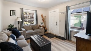 A living room with a sofa, ottoman, and a lamp. There is a door and a window with curtains. - Historic Drake Cottage - Walk to Downtown Bend - Drake Park Across the Street - Perfect for Couples (Bend)