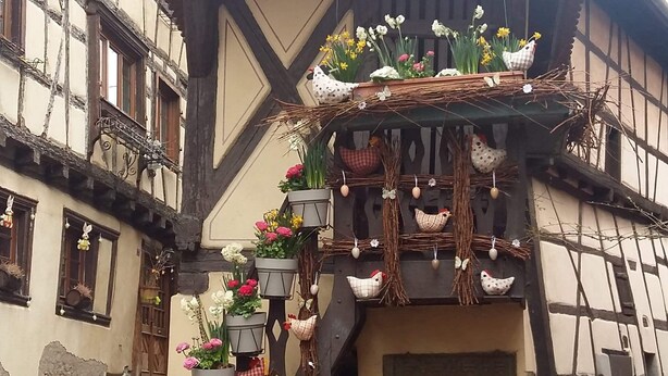 A traditional half-timbered building with a stone staircase leading to a balcony adorned with flowers and decorative chickens.