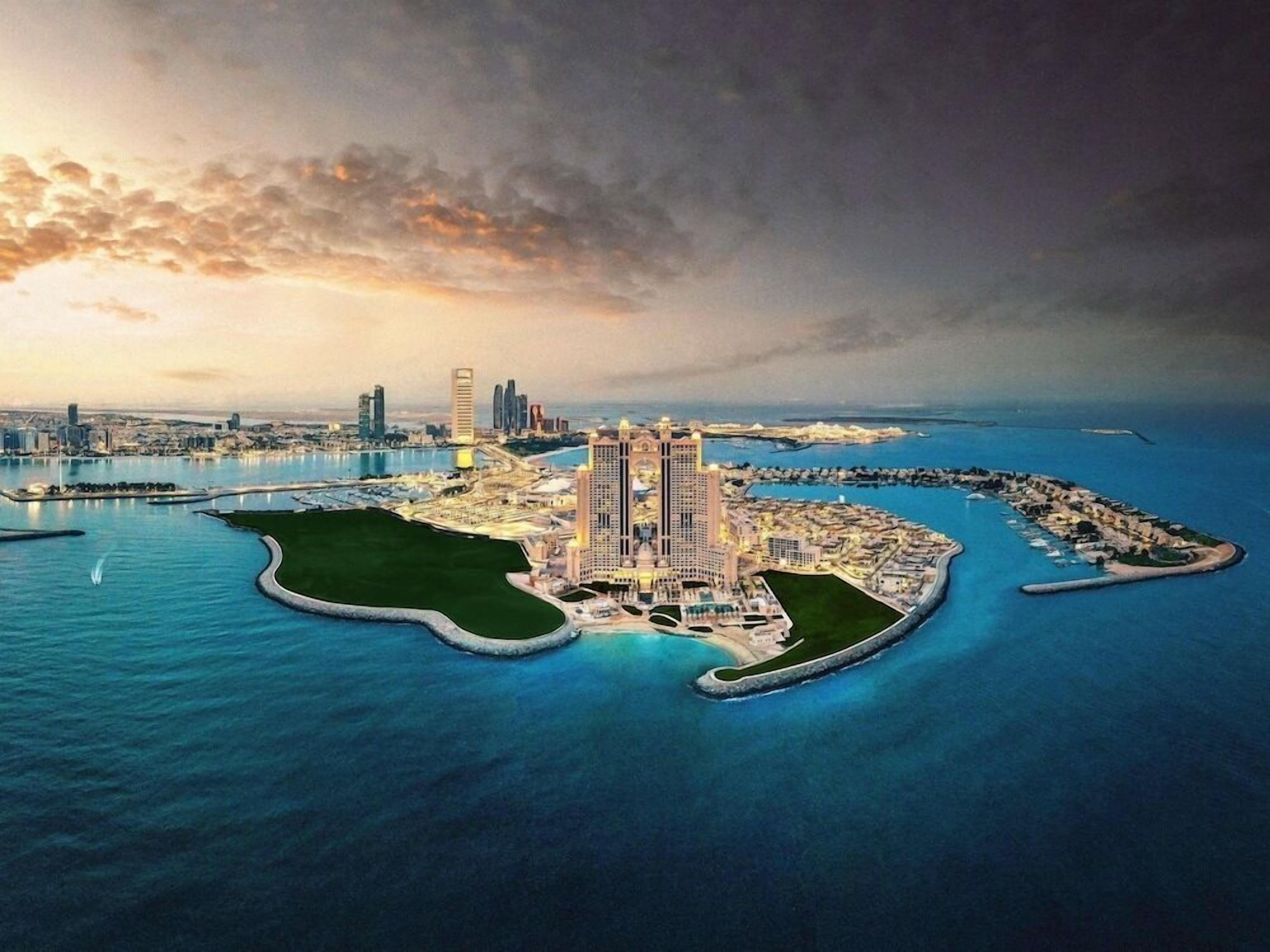A coastal cityscape with a prominent hotel featuring a central tower, surrounded by water and connected to the mainland by bridges.