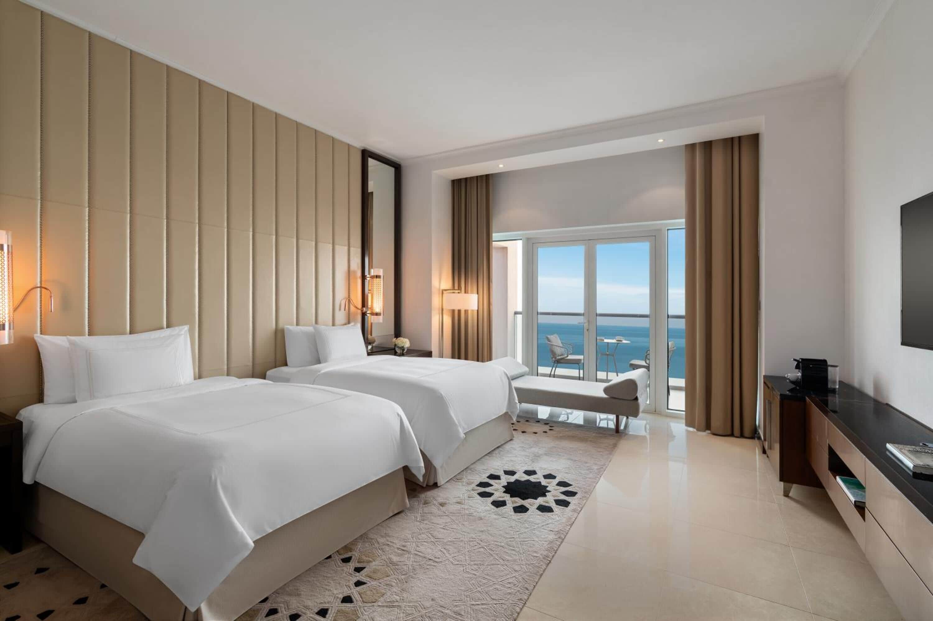 A hotel room with two beds, a large window with a view of the sea, a flat-screen TV, and a minibar.