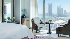 A hotel room with a large bed, two armchairs, a small table, a mirror, a view of a cityscape, and a desk with a lamp and decorative items. - Fairmont Marina Residences Abu Dhabi (Abu Dhabi)