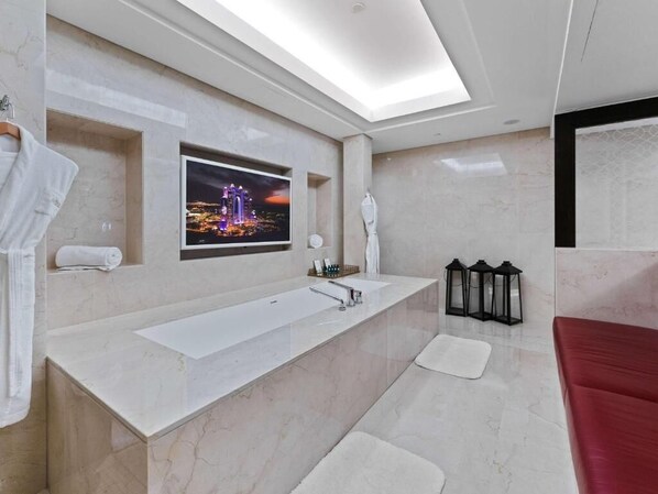 Une salle de bain moderne avec un grand miroir, deux lavabos, un téléviseur fixé au mur et un banc rouge.
