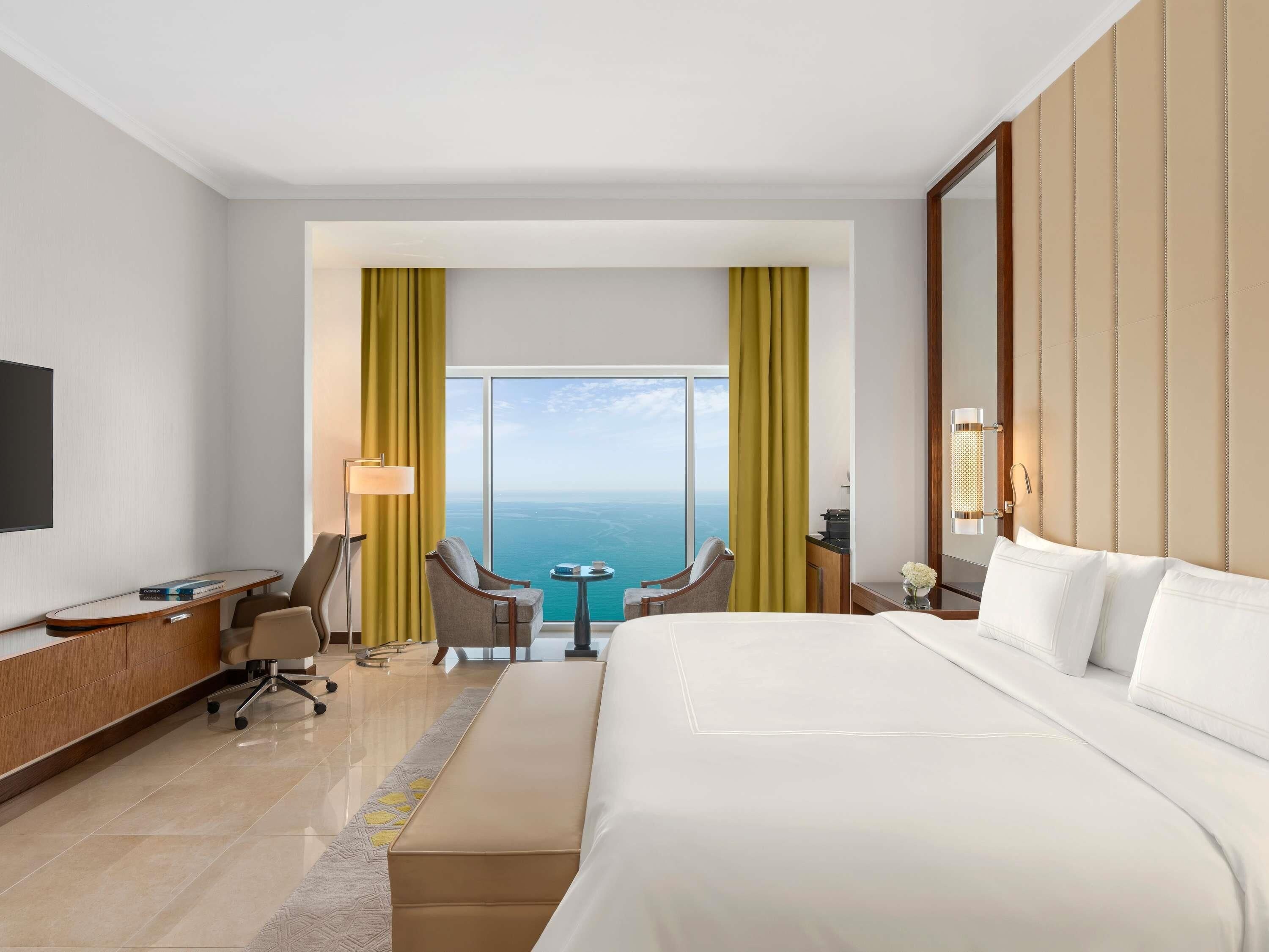 A hotel room with a large bed, a desk, two chairs, a small table, and a view of the ocean.