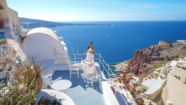Une terrasse avec une table et des chaises donnant sur la mer, une femme en robe blanche et une vue sur la côte.