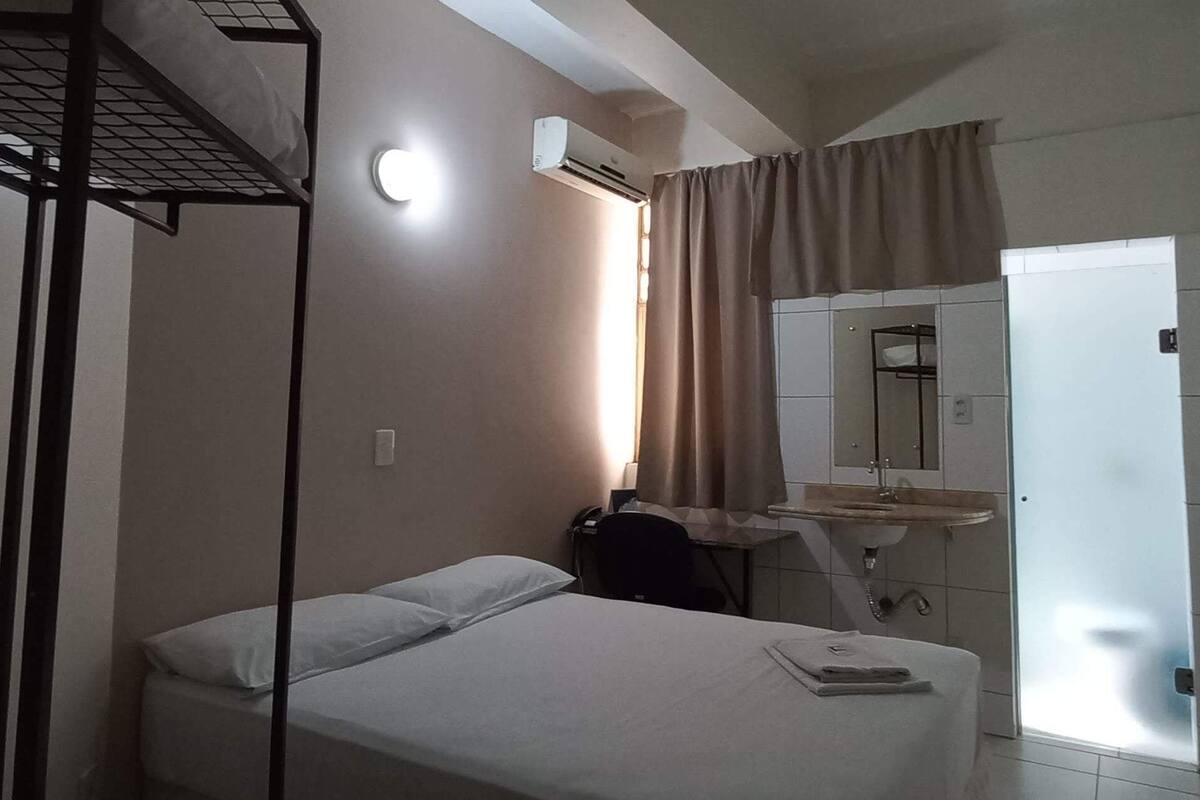 Une chambre d’hôtel avec un lit superposé, un bureau avec une chaise, une table de chevet avec un téléphone, un miroir et une salle de bain avec un lavabo et une douche.
