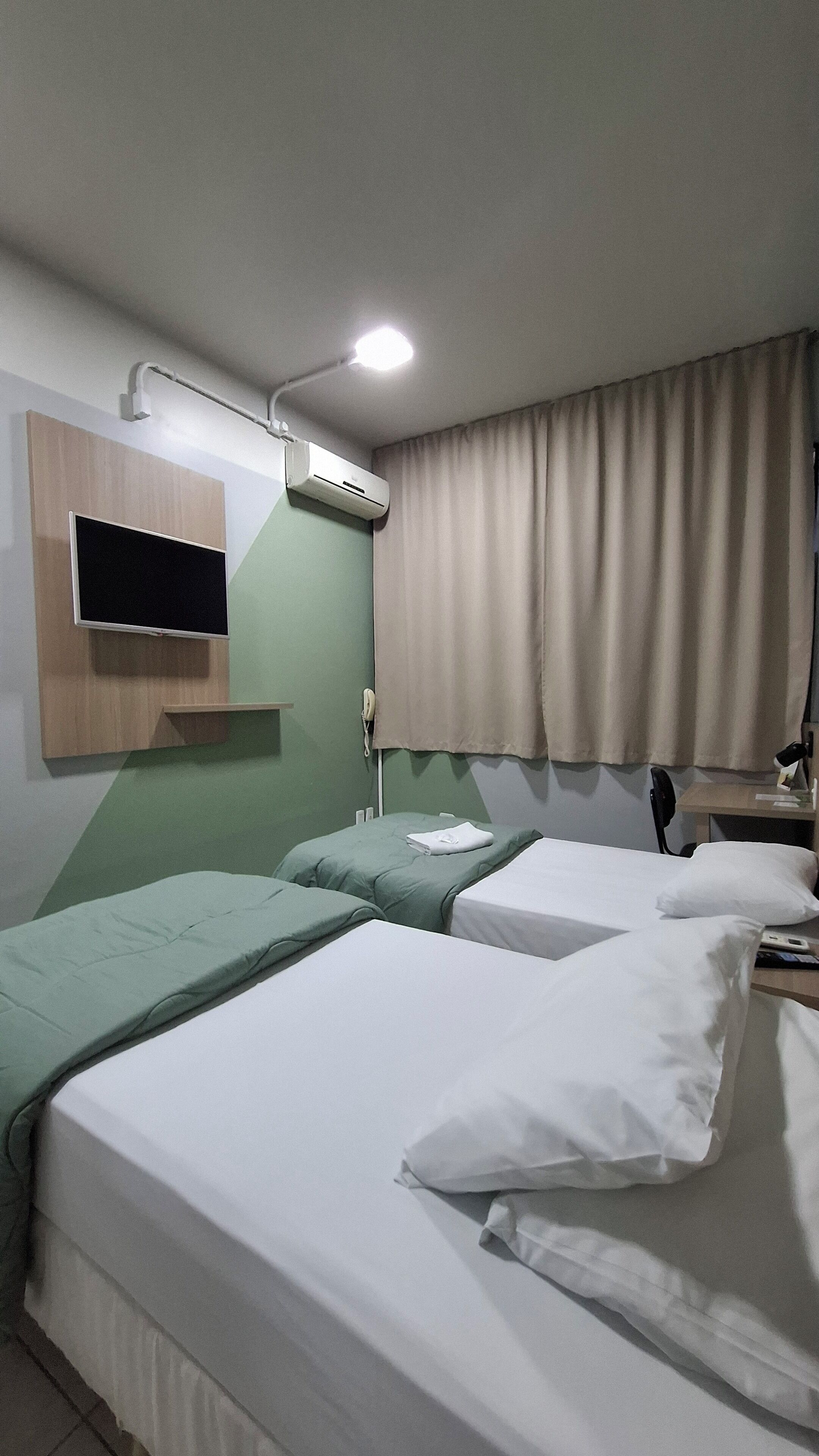 Habitación de hotel con dos camas, un televisor montado, equipo de aire acondicionado, una mesita de noche con lámpara y un teléfono.