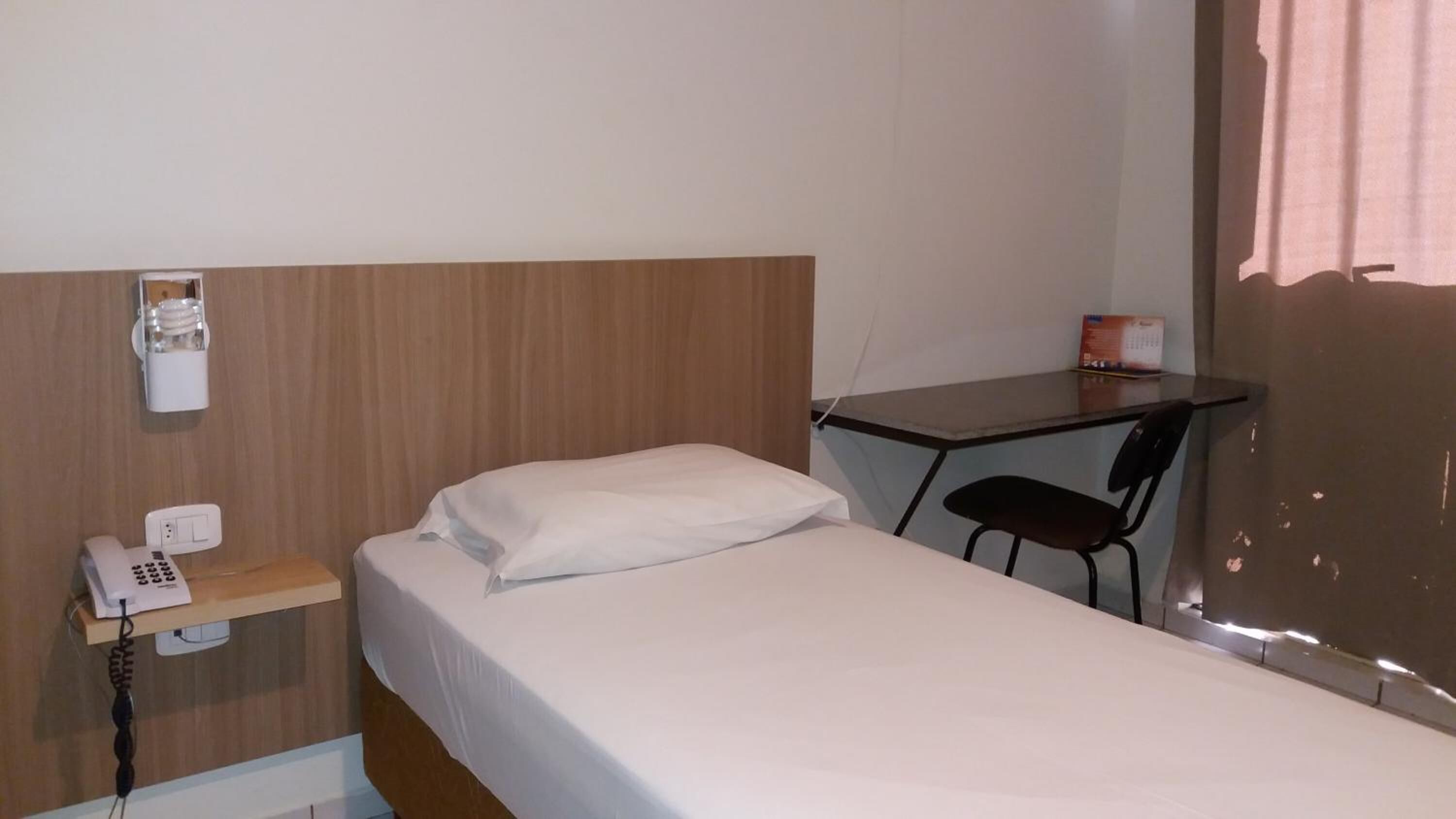 Una cama individual con un almohadón blanco y un cabecero de madera. Hay una mesita con una silla, un teléfono y un enchufe en la pared.