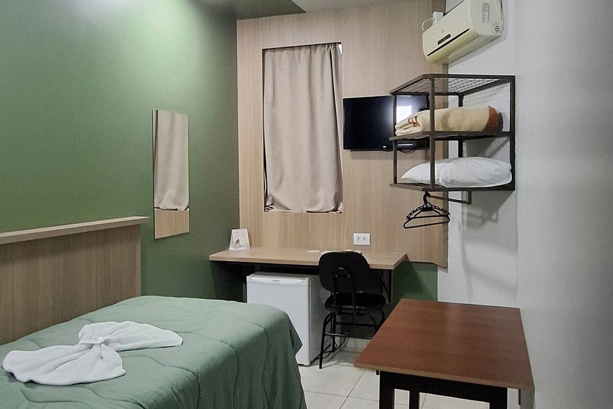 Une chambre d’hôtel de taille réduite, comprenant un lit simple, un bureau, une chaise, une petite table, un téléviseur et un climatiseur fixé au mur.