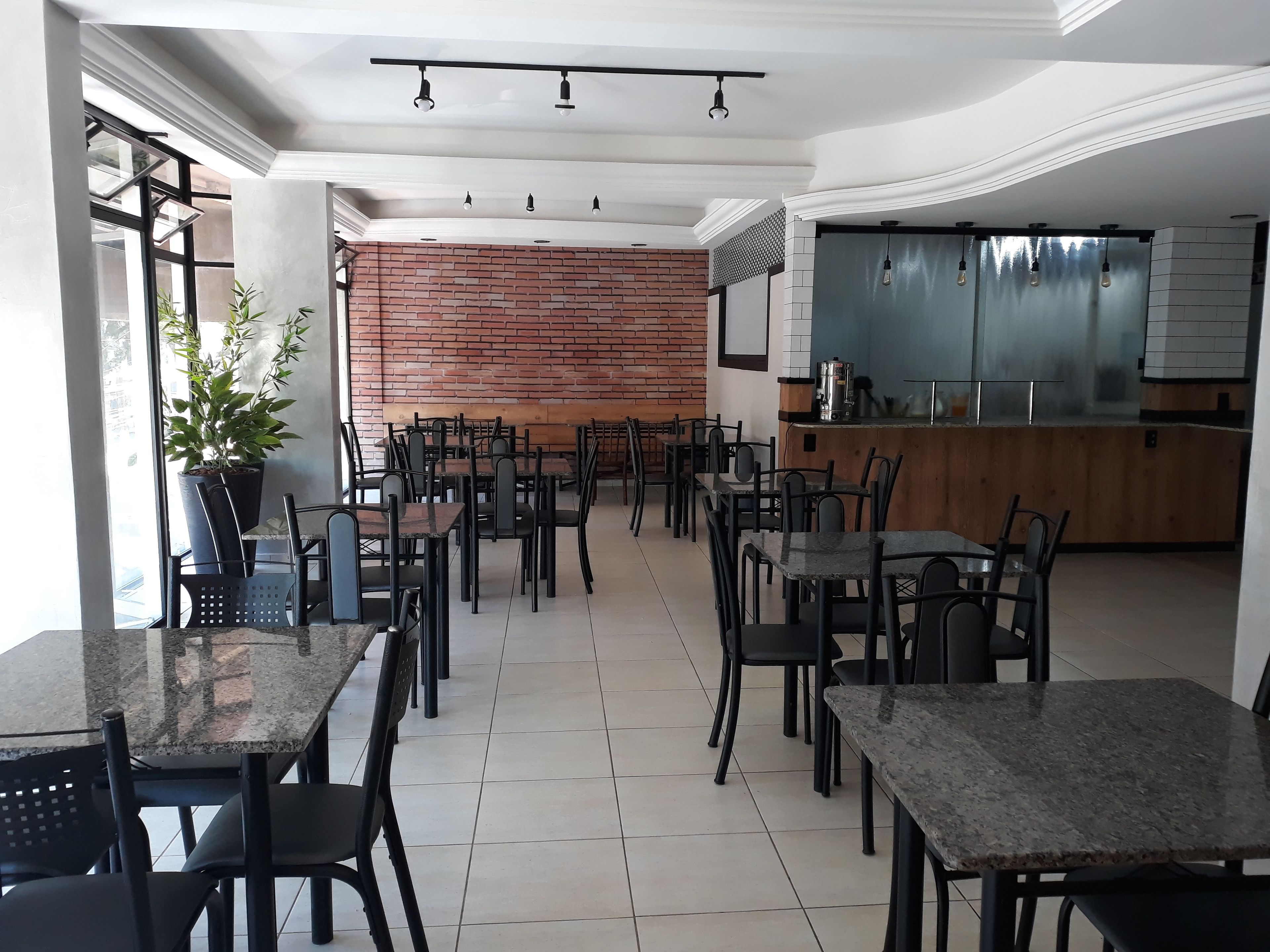 Uma sala de jantar moderna com várias mesas e cadeiras, uma parede de tijolos e uma área de cozinha com uma janela grande.