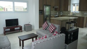 A modern living room with a grey sofa, a TV, and a small table. The kitchen features wooden cabinets, a refrigerator, and a granite countertop. - Casa Lila (Montecristi)