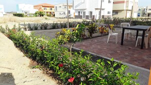 A patio area with a brick floor, surrounded by green plants and flowers. There are white plastic chairs and a table. In the background, there are multi-story buildings with a modern design. - Casa Lila (Montecristi)