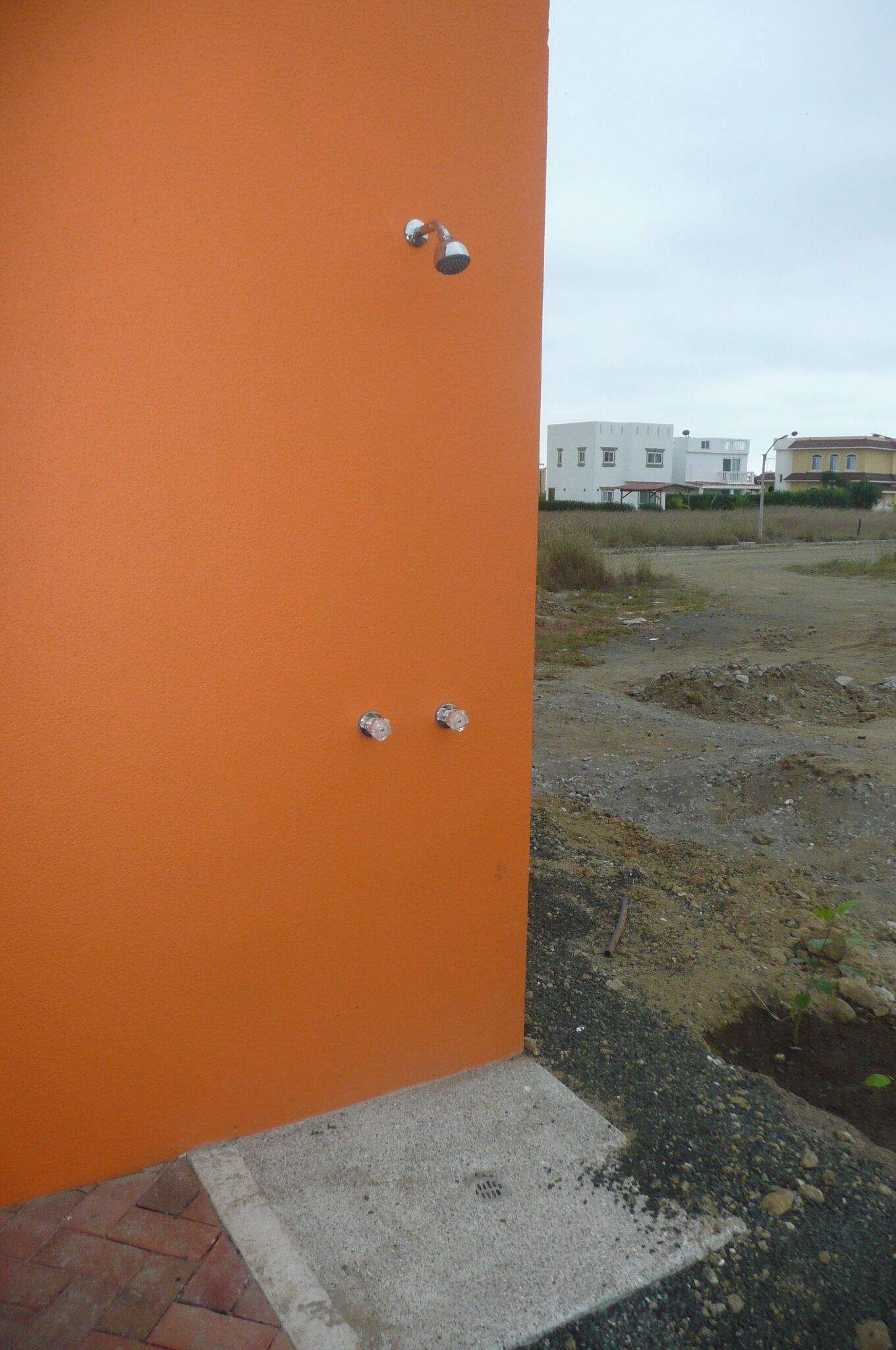 Esquina de una pared naranja con dos pomos metálicos y un escalón de hormigón que conduce a una zona de grava.