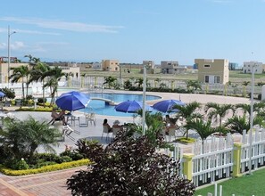 Un espace piscine d’un complexe hôtelier agrémenté de plusieurs parasols, de palmiers et offrant une vue dégagée sur les bâtiments environnants.