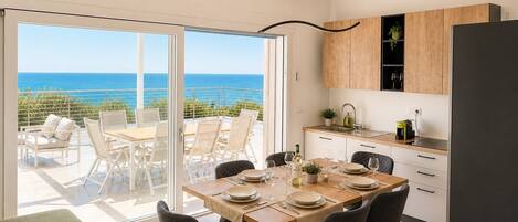 A modern kitchen with a view of the sea, a dining area with chairs and a table, and a large sliding glass door.