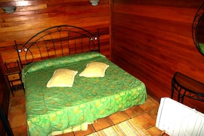 A single bed with a green bedspread and two pillows, a wooden wall, a small table with a chair, and a radiator. - Cabañas Valle Verde (Moya)