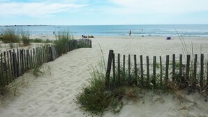 Une plage de sable bordée d’une clôture en bois, avec quelques plantes et une mer calme en arrière-plan.