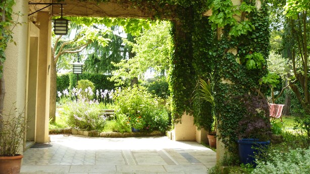 Un chemin de pierres serpente à travers un jardin luxuriant, bordé d’une pergola recouverte de plantes grimpantes vertes.