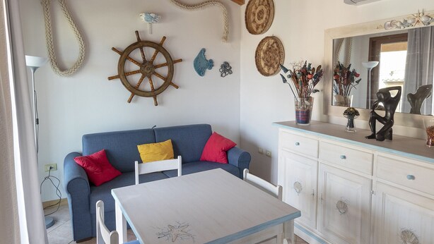 Una habitación acogedora con techo de madera, un sofá azul con cojines de colores, una mesa blanca con sillas azules y un mueble blanco con adornos.