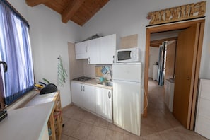 Une cuisine compacte avec des meubles blancs, un four à micro-ondes et un réfrigérateur. Il y a une fenêtre avec des rideaux bleus et un plafond en bois.