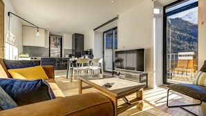 En moderne stue med sofa, sofabord og fjernsyn. Der er et køkkenområde med skabe og et spisebord. Værelset har store vinduer med udsigt til et bjerglandskab.