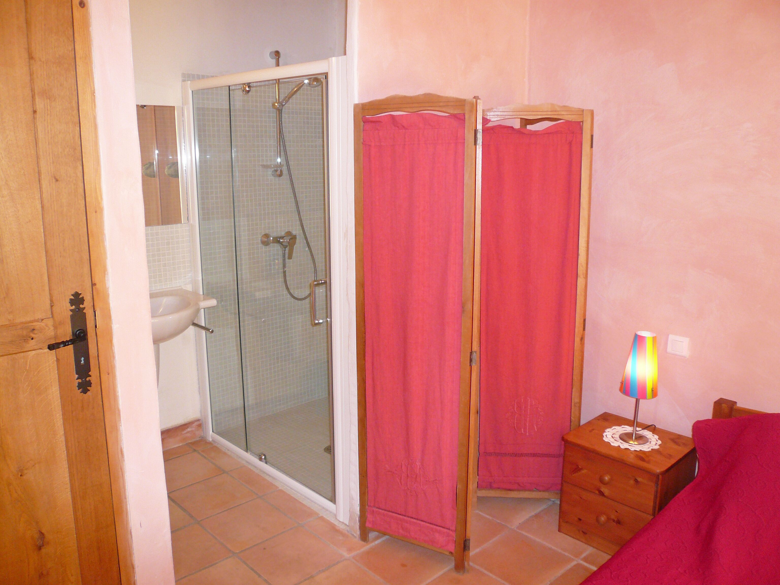 Un baño con ducha de vidrio, una puerta de madera, un lavabo y una mesita de noche con lámpara.