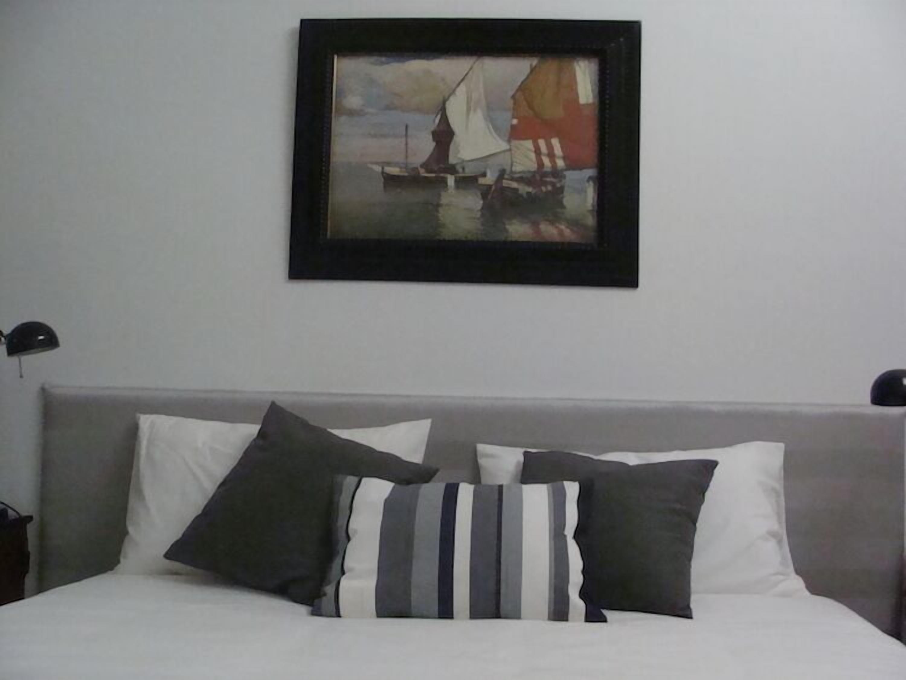Un lit bien fait, agrémenté d’oreillers blancs et à rayures, un tableau encadré représentant un voilier accroché au mur, et une simple lampe de chevet.