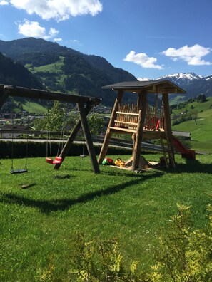 Ein hölzerner Spielplatz mit Schaukeln und Rutsche vor sanften, grünen Hügeln und blauem Himmel.