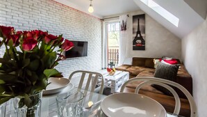A dining area with a glass table, white plates, and a vase of red roses. There is a white brick wall and a sofa with a red cushion in the background. - Apartamenty hoteLOVE pod Debami (Karpacz)