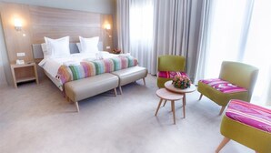 Une chambre d’hôtel avec un grand lit, une banquette, deux fauteuils, une petite table ronde et une table de chevet avec une lampe.