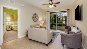 A living room with a beige sofa, a small armchair, a coffee table, and a TV mounted on the wall. There is a clock on the wall and a ceiling fan above the sofa. - Magnolia Style Condo at Windsor Hills with Disney Theme Bunk Room (Kissimmee)