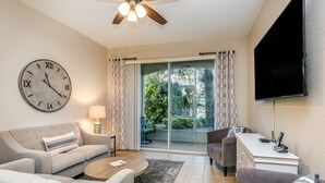 A living room with a sofa, coffee table, and a TV. There's a large clock on the wall and a ceiling fan. - Magnolia Style Condo at Windsor Hills with Disney Theme Bunk Room (Kissimmee)