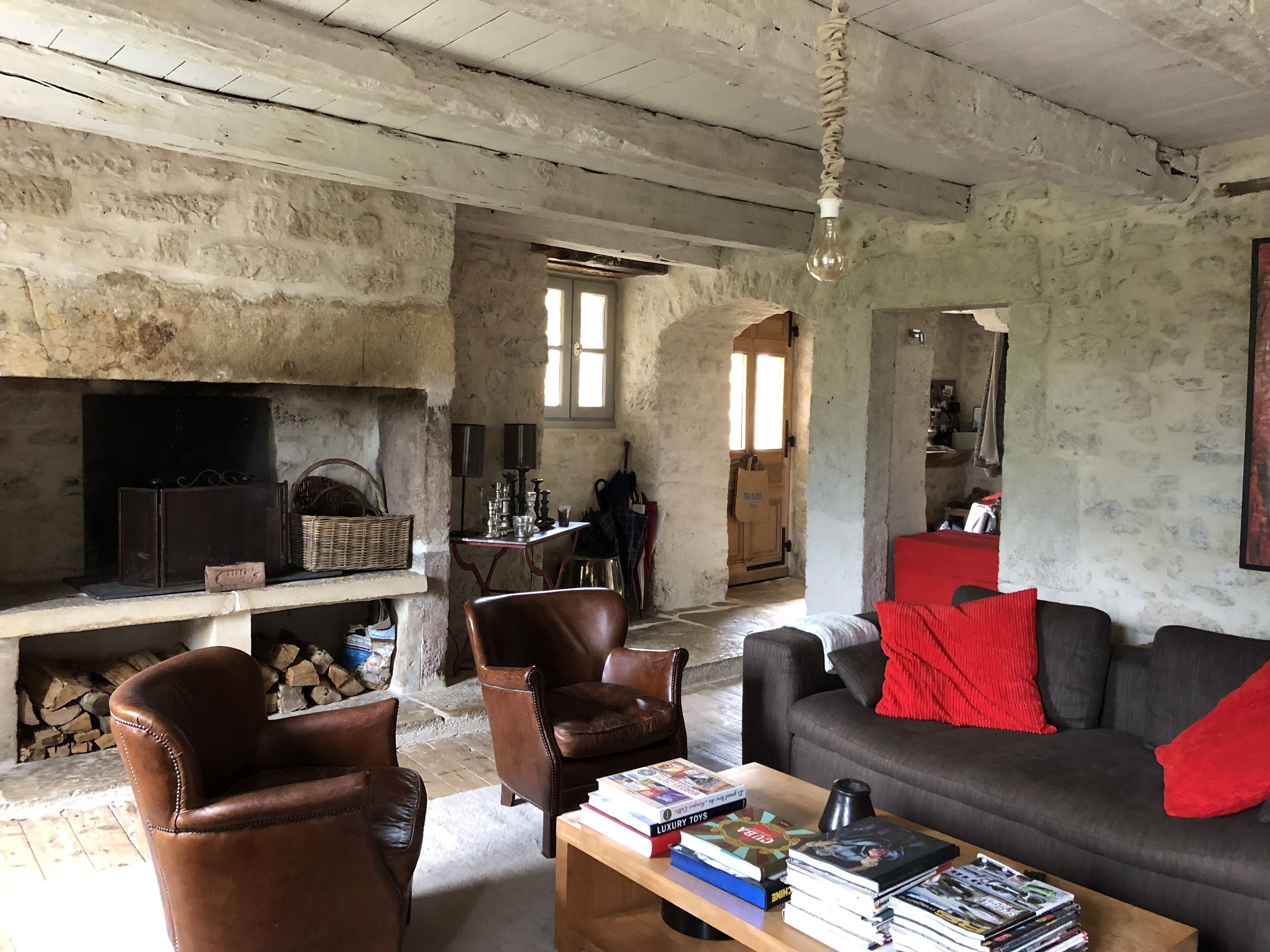 Un salon rustique avec une cheminée, des fauteuils en cuir, un canapé avec des coussins rouges et une table basse sur laquelle sont posés des livres et des magazines.