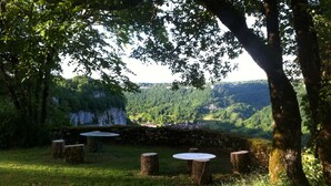 A rustic outdoor seating area with stone tables and benches, surrounded by lush greenery and overlooking a valley with a distant village. - Beautiful view of Baume les Messieurs (Baume les Messieurs)